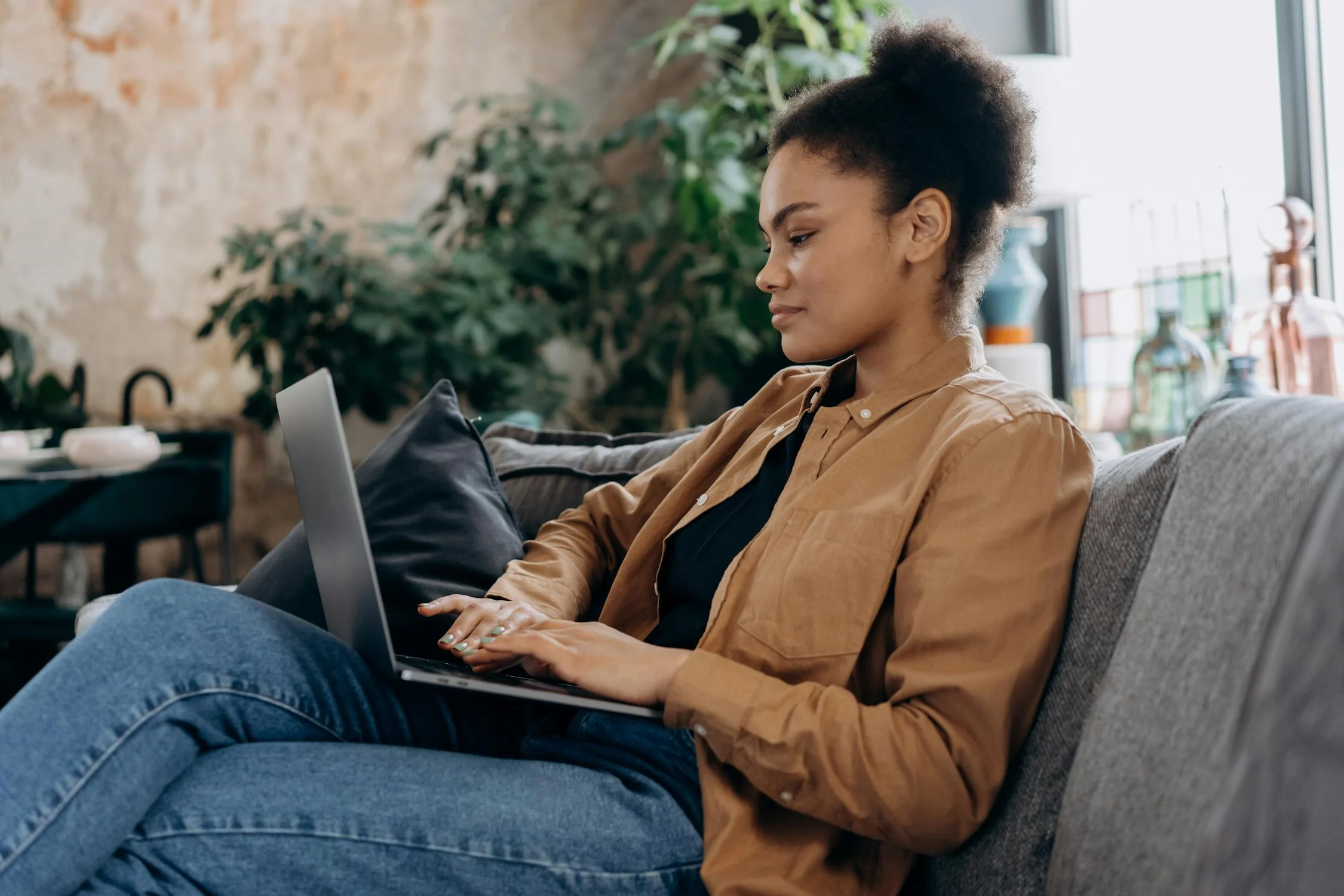 A young woman with curly hair tied up in a bun is sitting on a gray couch, working on a laptop. She is wearing a brown jacket over a black top and blue jeans. The background features indoor plants and a cozy, modern living space with a rustic wall.