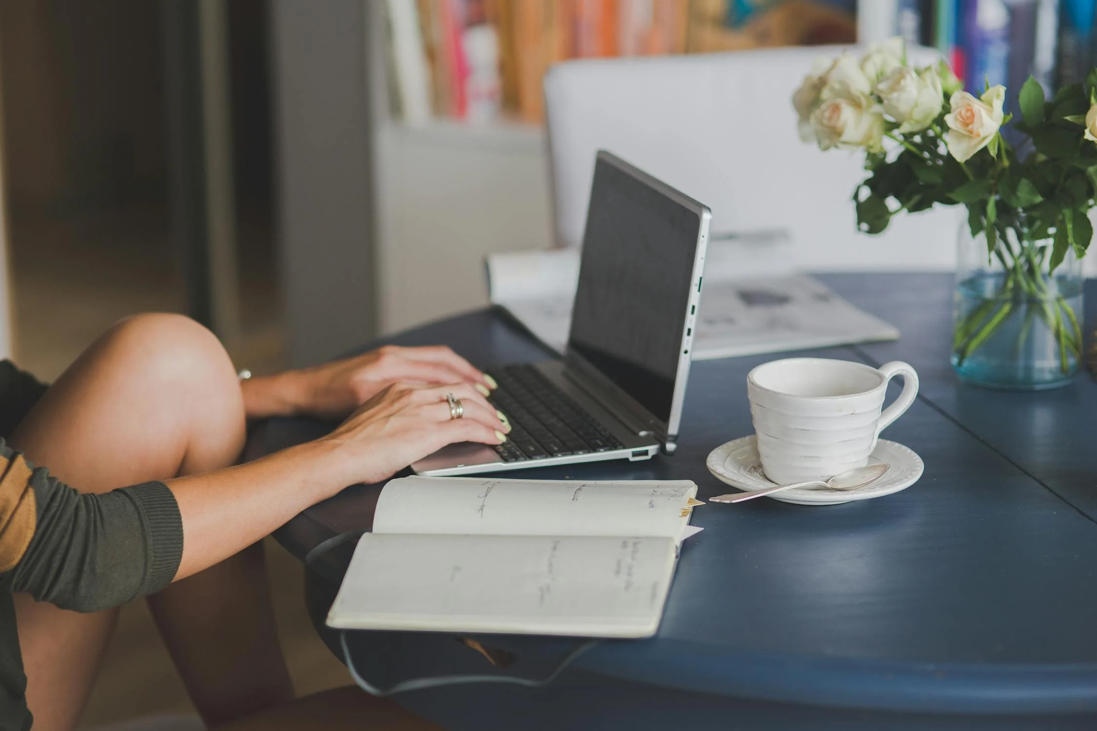 A woman working on a laptop at a dark blue table with a notebook, a white coffee cup on a saucer, and a vase of white roses in the background.