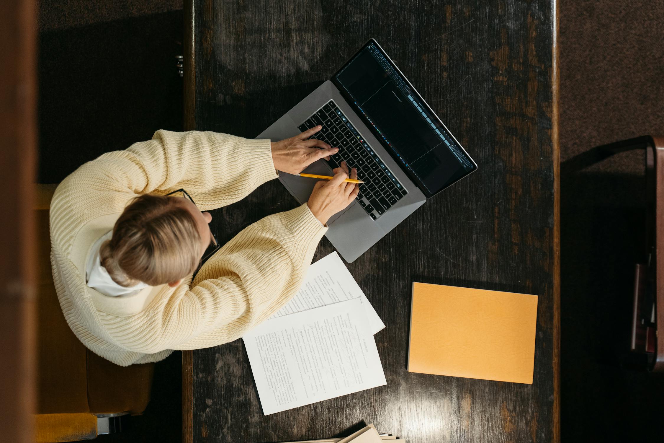 Top-down view of a person in a cream-colored sweater working on a laptop at a dark wooden table, with documents, a yellow folder, and a pencil in hand.