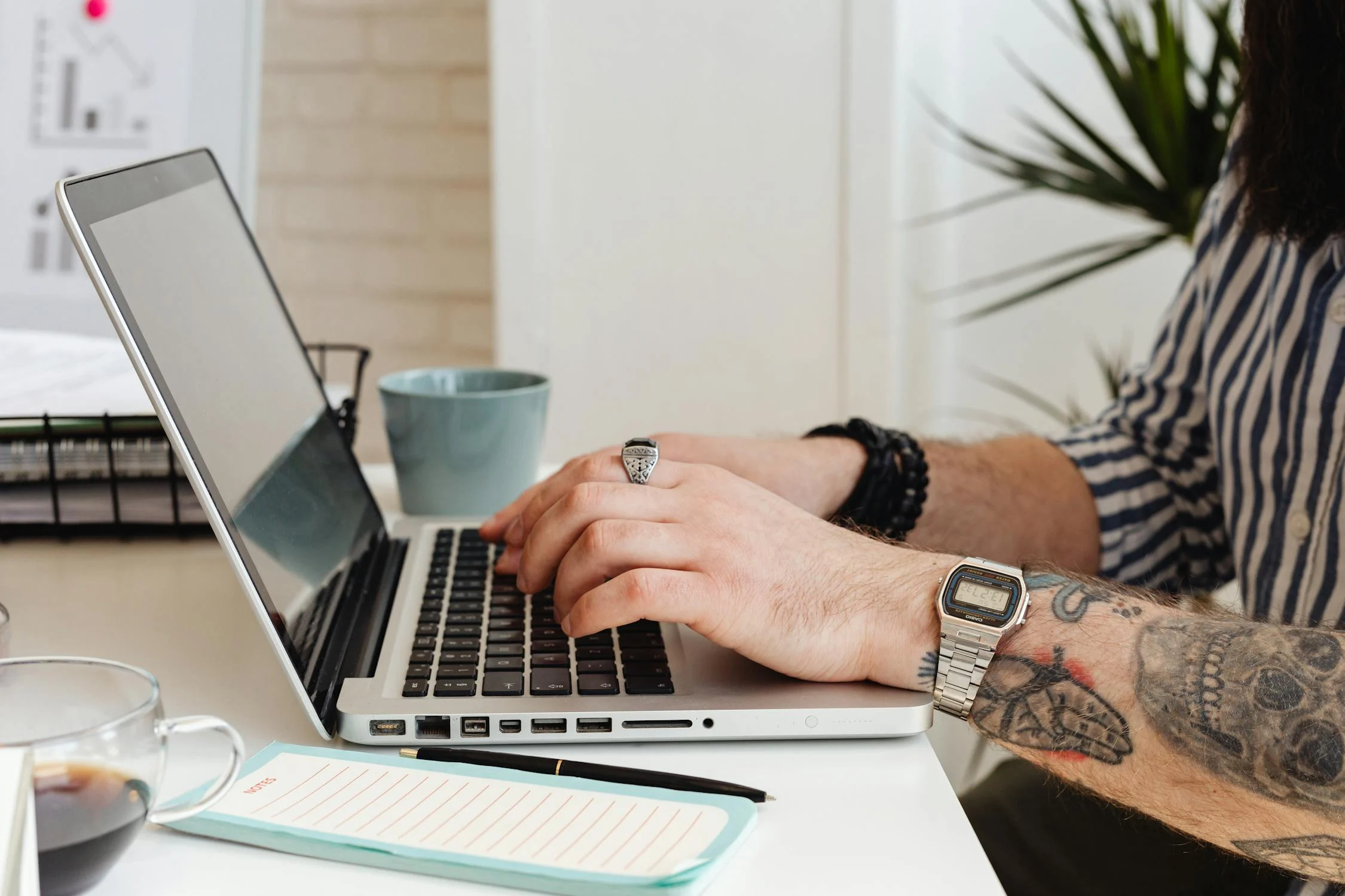 A person with tattoos and accessories typing on a laptop at a white desk, with a notepad, pen, coffee cup, and plant in the background.